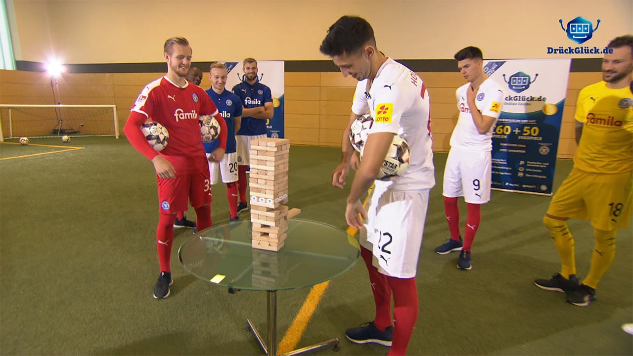 Fußballspieler spielen Jenga in der Halle, DrückGlück.de-Branding im Hintergrund Fußballspieler spielen Jenga in der Halle, DrückGlück.de-Branding im Hintergrund
